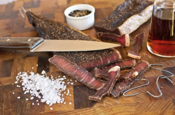 A cutting board with Biltong Meat Spiced U-Dry, salt, and a knife.