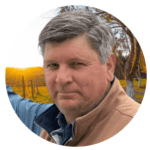 A man with gray hair wearing a brown vest and blue shirt is standing outdoors with trees and a sunset in the background, ready to offer expert tips on cooking meat perfectly every time.